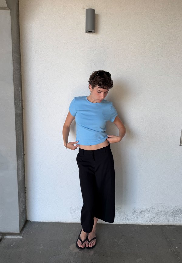 Personne avec des cheveux courts et bouclés portant un t-shirt bleu, un pantalon noir à jambes larges et des tongs, debout contre un mur blanc à l’intérieur.