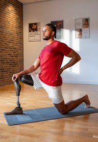 Prothese van een been met een afwerking van carbonvezel, een rood sportshirt en een witte short. Persoon knielt op een blauwe oefeningmat in een studio.
