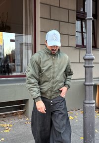 Veste bomber vert olive avec fermeture �éclair à l'avant et poches, portée avec un pantalon large en denim foncé. Porté avec une casquette de baseball bleu clair.