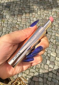 KIKO One Magic Touch styl kiss w srebrnej tubie ze złotymi akcentami, prezentującym różową szminkę, trzymanym na tle brukowanej ulicy.