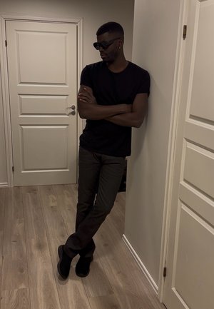 Hombre con gafas de sol negras, camiseta negra y pantalones oscuros apoyado contra una pared con los brazos cruzados en una habitación con suelo de madera y puertas blancas.