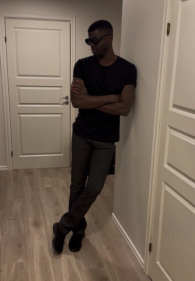 Hombre con gafas de sol negras, camiseta negra y pantalones oscuros apoyado contra una pared con los brazos cruzados en una habitación con suelo de madera y puertas blancas.