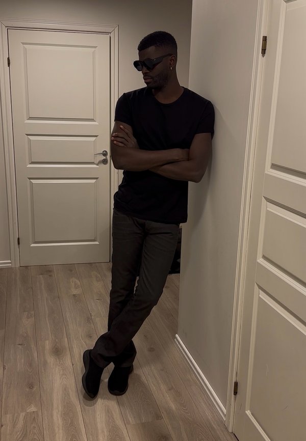 Homme portant des lunettes de soleil noires, un t-shirt noir et un pantalon foncé, appuyé contre un mur les bras croisés dans une pièce avec un sol en bois et des portes blanches.