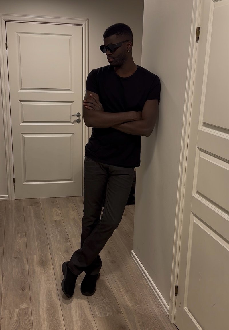 Homme portant des lunettes de soleil noires, un t-shirt noir et un pantalon foncé, appuyé contre un mur les bras croisés dans une pièce avec un sol en bois et des portes blanches.