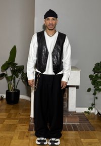 Uomo che indossa un gilet in pelle nero sopra un maglione bianco, pantaloni neri a gamba larga e sandali bianchi e neri, con una catena d'oro e un berretto.