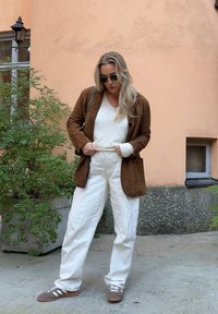 Brun suedjakke over en hvit genser, parret med hvite bukser og brune sneakers, med en strukturert overflate og en veske med kjede-accent.