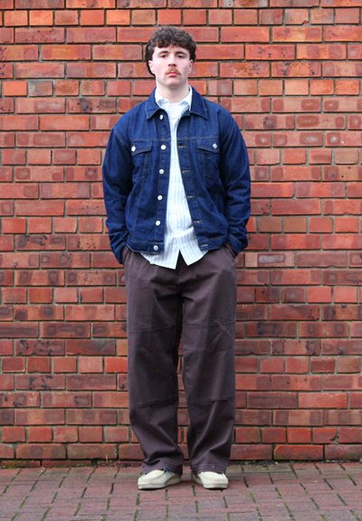 Chaqueta de mezclilla sobre una camisa blanca con rayas azules, combinada con pantalones holgados marrones y zapatos de color claro, frente a un fondo de pared de ladrillo.