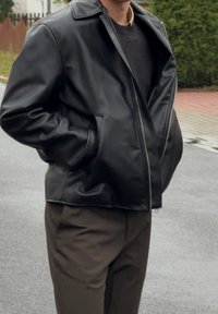 Zwarte leren bomberjack met een rits aan de voorkant, brede revers en zijzakken. Gedragen over een donkere trui, in combinatie met bruine broek.