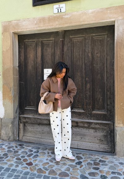 Mujer con chaqueta marrón y pantalones blancos con lunares sostiene una bolsa beige mientras está de pie en una calle empedrada frente a una gran puerta de madera oscura.