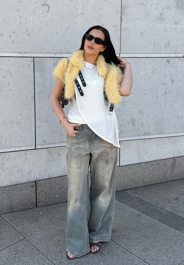 Femme aux longs cheveux foncés portant des lunettes de soleil noires, un haut blanc avec un gilet en fourrure jaune, un pantalon large gris et des chaussures ouvertes, debout contre un mur de pierre.