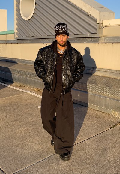 Chaqueta bomber acolchada negra sobre una sudadera con capucha negra, combinada con pantalones negros anchos y botas negras gruesas. Los accesorios incluyen collares en capas y un gorro de lana.