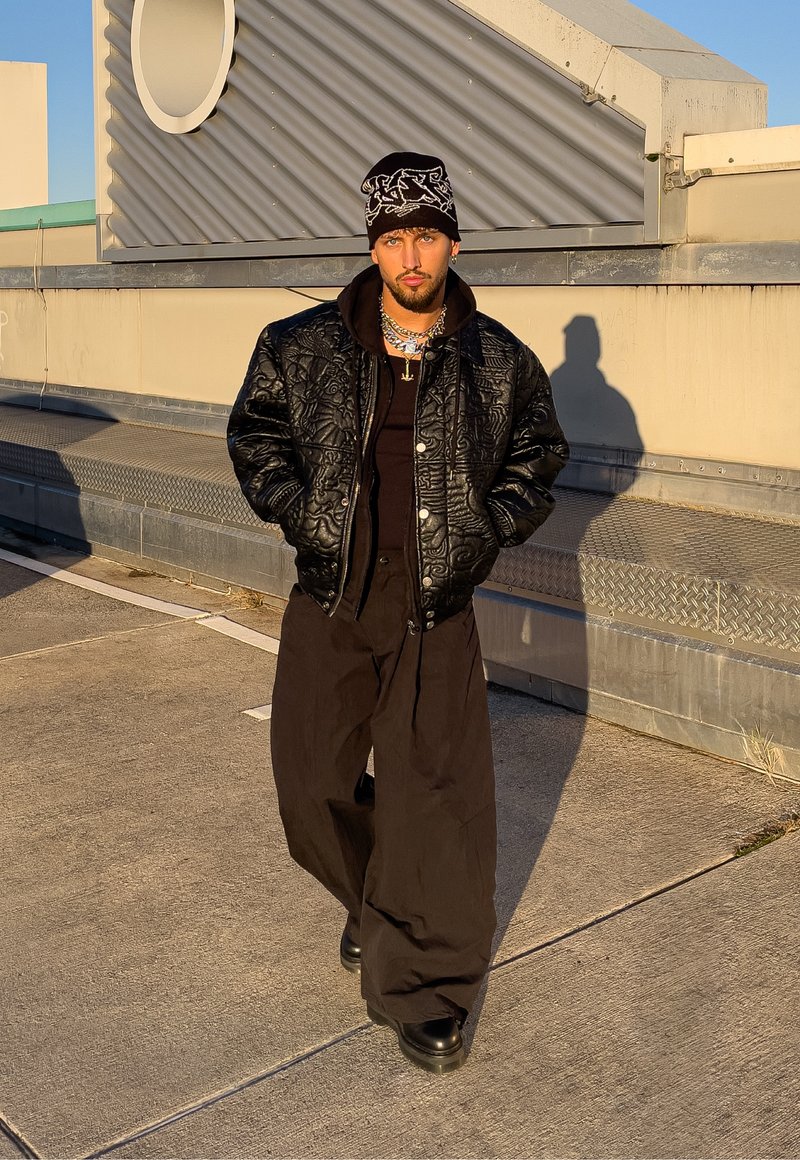 Bomber jacket trapuntato nero sopra una felpa con cappuccio nera, abbinata a pantaloni neri larghi e stivali neri robusti. Accessori includono collane stratificate e un berretto.