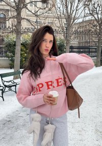 Jeune femme en pull rose tenant une tasse de café et un sac marron, debout sur un sentier enneigé avec des arbres dénudés et des bancs à proximité.