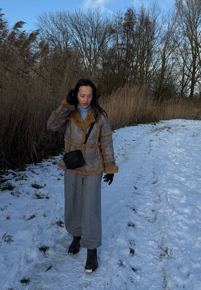Mujer con abrigo de invierno y guantes caminando por un camino nevado junto a juncos altos y secos bajo árboles desnudos y cielo azul.