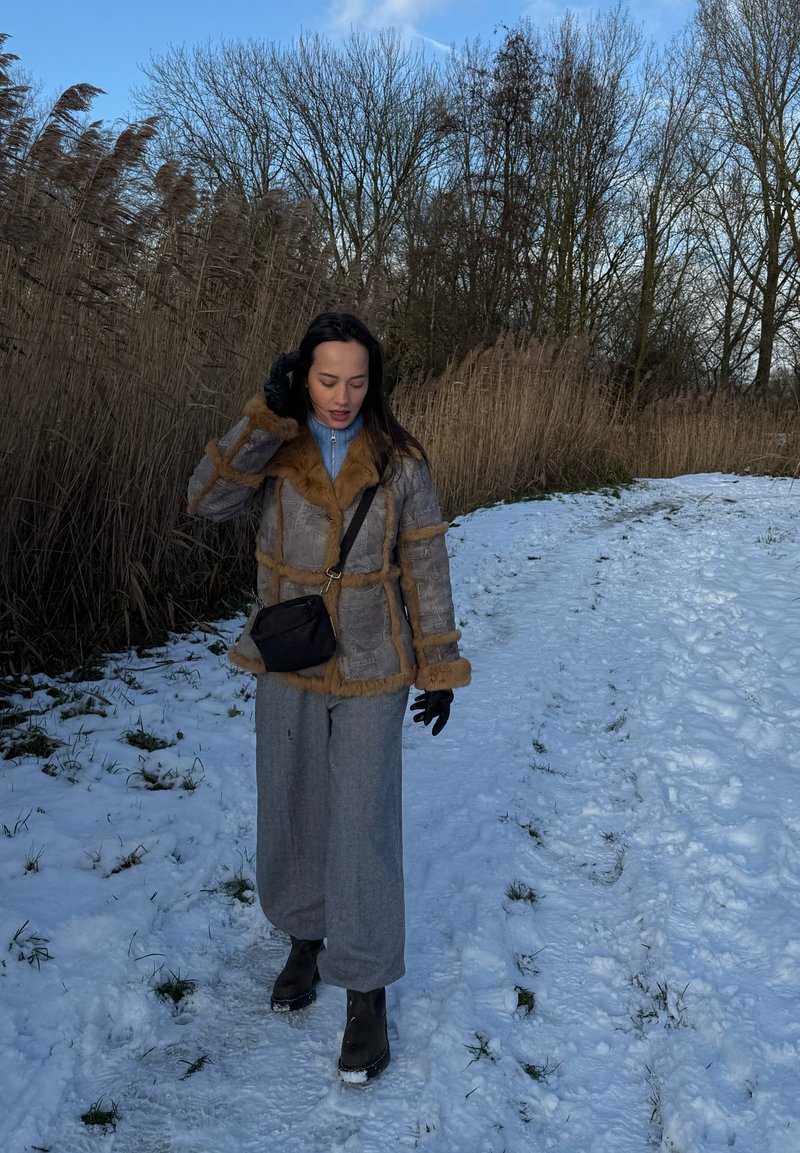 Femme en manteau d'hiver et gants marchant sur un sentier enneigé à côté de hautes roseaux secs sous des arbres dénudés et un ciel bleu.