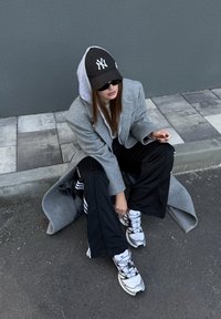 Manteau long gris sur un sweatshirt blanc, lunettes de soleil noires, pantalon noir avec des rayures blanches, et chaussures de sport blanches avec des accents noirs.