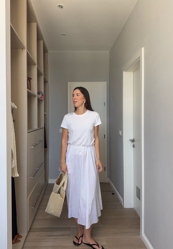 Frau in weißem T-Shirt und Rock hält eine geflochtene Tasche und steht in einem schmalen Flur mit beigen eingebauten Regalen und geschlossenen Türen.