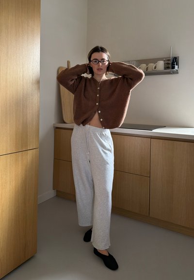 Persona ajustándose las gafas, vestida con un cárdigan marrón, pantalones deportivos gris claro y zapatos negros, de pie en una cocina moderna de madera.