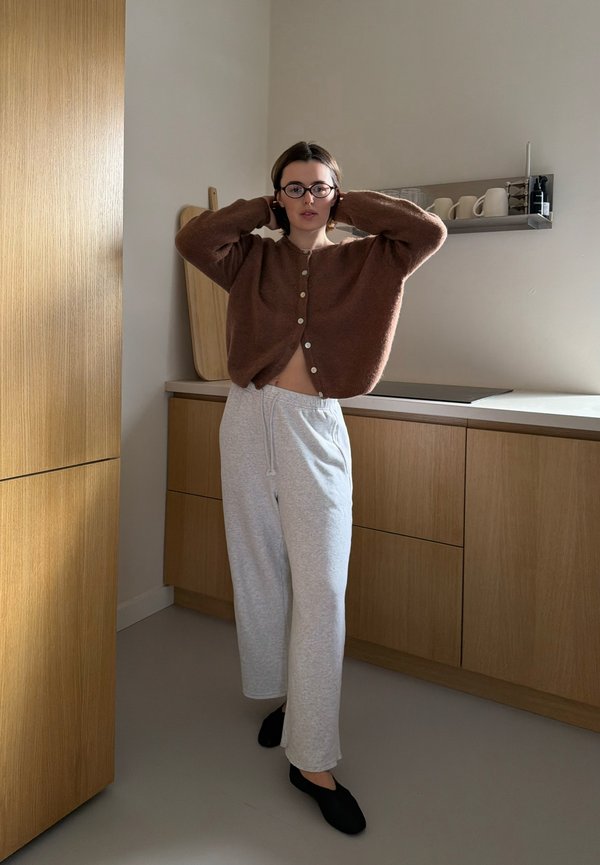 Osoba koja podešava naočale, nosi smeđi kardigan, svijetlosive trenirke i crne cipele, stojeći u modernoj drvenoj kuhinji.