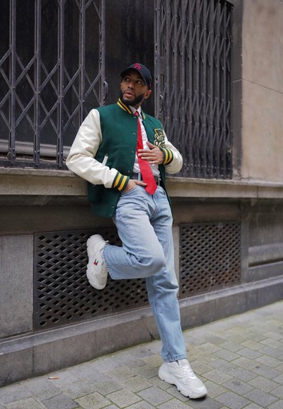 Chaqueta varsity verde y crema con puños acanalados, corbata roja, jeans azules claros y zapatillas blancas. Apoyado contra una pared texturizada.