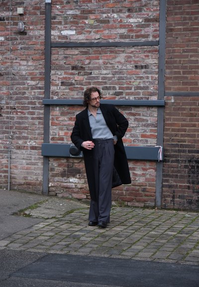 Abrigo largo negro sobre una camisa gris con cuello y pantalones grises de pierna ancha. Lleva puesto unos zapatos negros; de pie contra una pared de ladrillo.