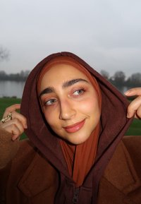 Brun hættetrøje lagdelt over et rustfarvet tørklæde; glat tekstur, afslappet pasform og neutral makeup med naturlige træk.