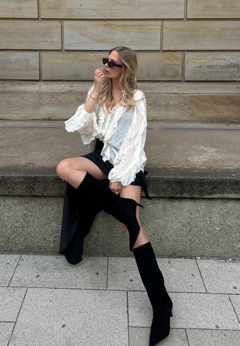 Blouse blanche à volants, mini jupe noire et bottes en velours noires montant jusqu'aux cuisses. Une personne assise sur une surface en pierre avec un arrière-plan texturé.