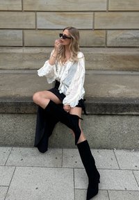 Blouse blanche à volants, mini jupe noire et bottes en velours noires montant jusqu'aux cuisses. Une personne assise sur une surface en pierre avec un arrière-plan texturé.