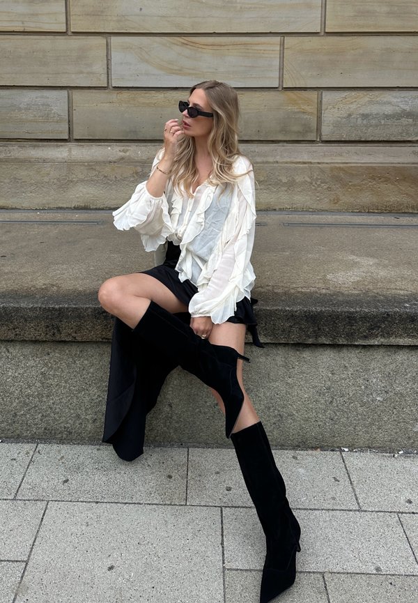 Blusa branca com babados, saia mini preta e botas altas de veludo pretas. Pessoa sentada sobre uma superfície de pedra com um fundo texturizado.