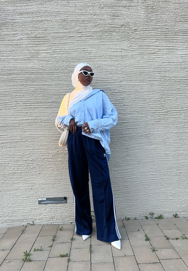 Lyseblå oversized skjorte med knapper, marineblå joggebukser med hvite striper, hvite solbriller, hvit håndväske og hvite spisse sko.