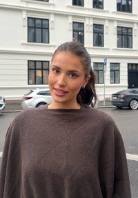 Giovane donna con capelli scuri raccolti, indossa un maglione marrone, in piedi su una strada con auto parcheggiate e finestre di un edificio bianco dietro di lei.