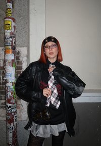 Veste en cuir noire, chemise noire avec un motif rouge, cravate à carreaux, jupe grise, collants noirs. Les accessoires comprennent un bandeau noir et un sac.