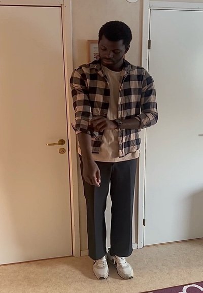 Hombre remangándose la camisa a cuadros en blanco y negro en el interior, de pie entre dos puertas cerradas sobre un suelo beige.