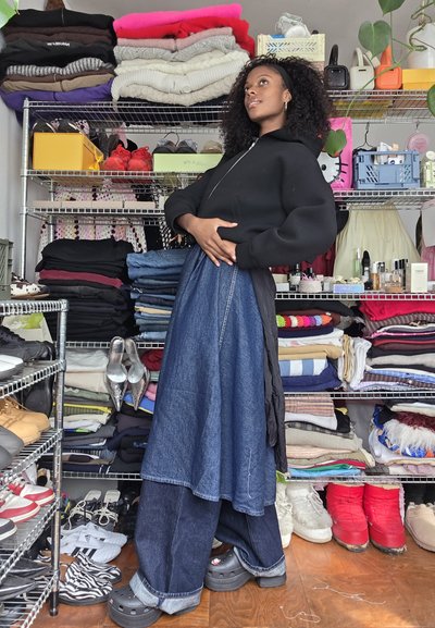 Sudadera negra con cremallera, combinada con una falda de mezclilla larga sobre unos vaqueros de pierna ancha. Lleva Crocs negros en los pies y hay zapatos plateados en el estante. Ropa de colores doblada en el fondo.
