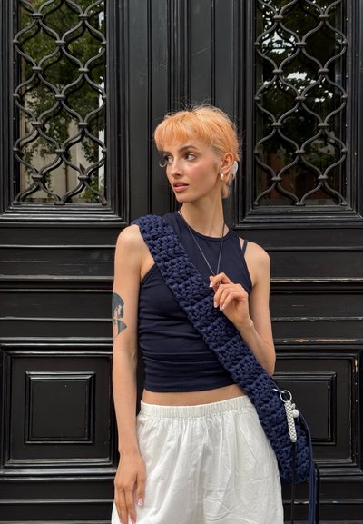 Bolso de hombro de ganchillo azul marino con una correa gruesa y texturizada. La modelo lleva una blusa ajustada negra y pantalones blancos holgados, de pie frente a una puerta negra ornamentada.