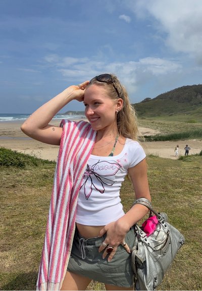 Joven mujer con gafas de sol y toalla a rayas sobre el hombro sonríe en la playa, sosteniendo un bolso plateado con un objeto rosa en su interior.