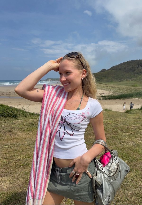 Junge Frau mit Sonnenbrille und gestreiftem Handtuch über der Schulter lächelt am Strand und hält eine silberne Handtasche mit einem pinkfarbenen Gegenstand darin.