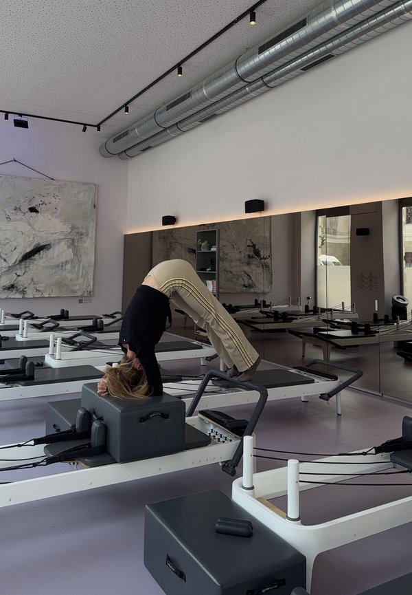 Osoba vykonávajúca Pilates cvičenie na reformer stroji v jasnom štúdiu s veľkými zrkadlami a moderným dekorom.