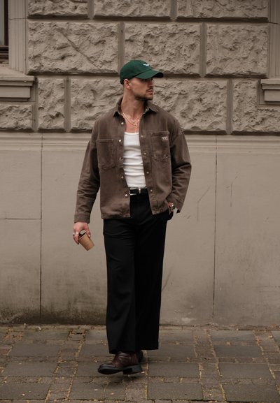 Camisa de pana marrón sobre una camiseta blanca de tirantes, pantalones anchos negros y zapatos de cuero oscuros. Sosteniendo un objeto cilíndrico marrón. Gorro verde.