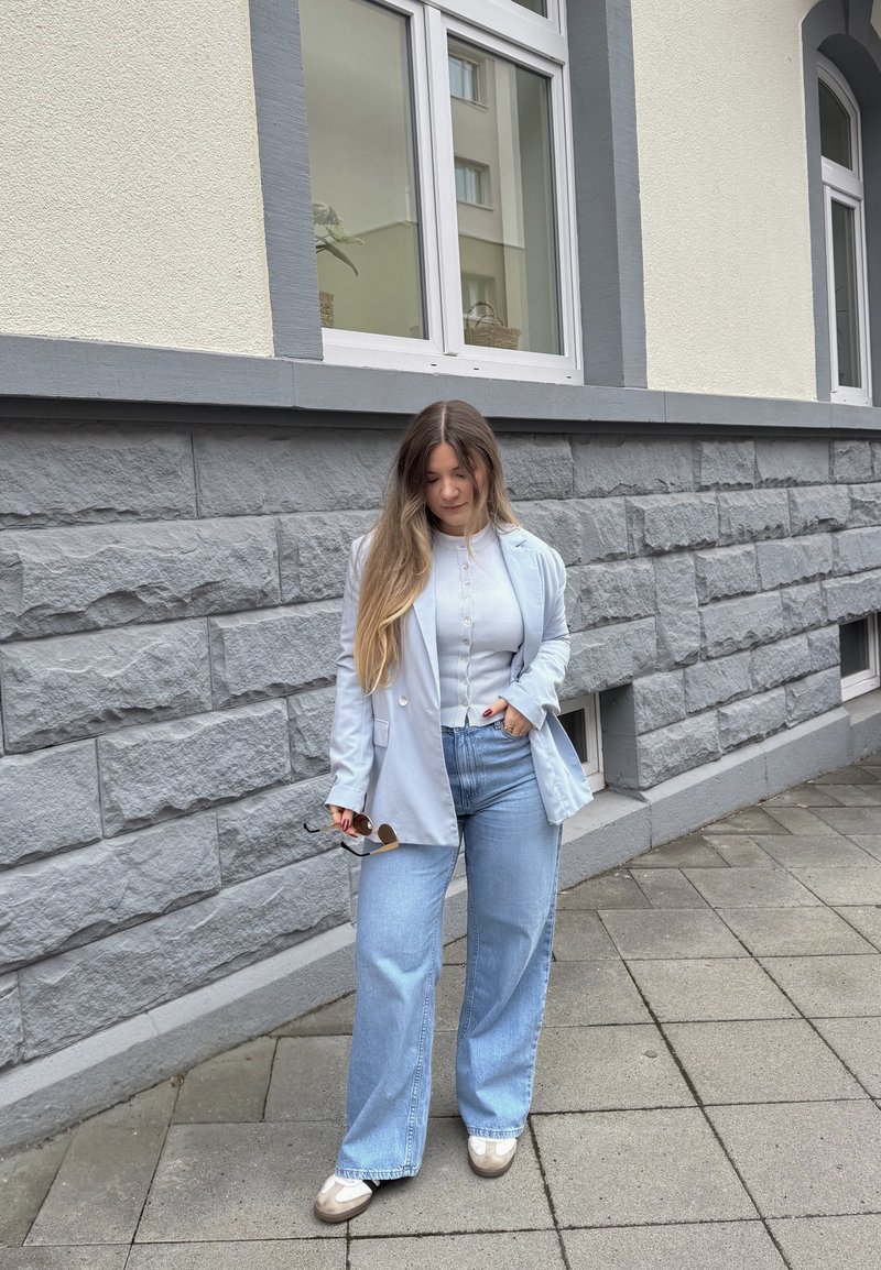 Femme aux longs cheveux portant un blazer bleu clair, un haut blanc, un jean large, tenant des lunettes de soleil, debout sur un trottoir pavé près d'un mur en pierre grise.