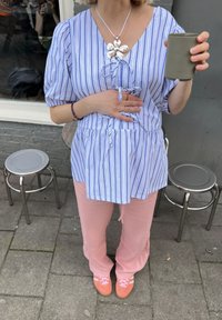 Ριγέ μπλουζάκι σε μπλε και λευκό χρώμα με λεπτομέρεια σε ζωνάκι, συνδυασμένο με ροζ παντελόνια και πορτοκαλί παπούτσια με λευκές ρίγες. Κρατάει ένα πράσινο φλιτζάνι.