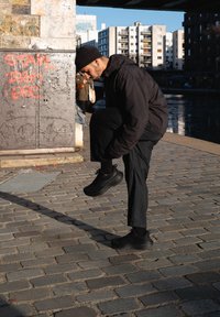 Homme en tenue noire et bonnet nouant son lacet sur un chemin pavé près d’un canal et d’un mur couvert de graffiti en milieu urbain.