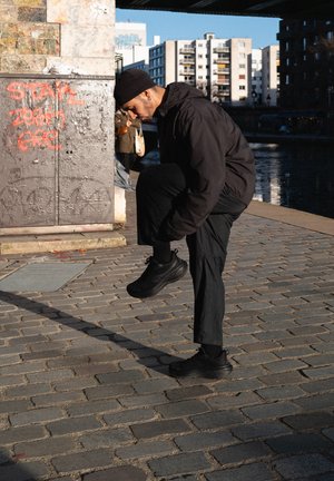 Hombre con atuendo negro y gorro atándose los cordones de los zapatos en un camino de adoquines cerca de un canal y una pared cubierta de graffiti en un entorno urbano.
