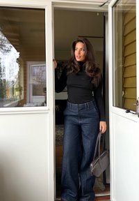 Kobieta w czarnej, długorękawowej bluzce i szerokich dżinsach, trzymająca małą brązową torebkę, stojąca w progu drzwi z jedną ręką opartą o framugę.