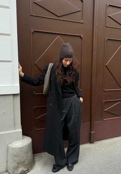 Abrigo negro de doble botonadura sobre una blusa negra, combinado con pantalones de pata de elefante con rayas. Lleva un gorro de punto oscuro y una bolsa trenzada. Fondo de puerta marrón.