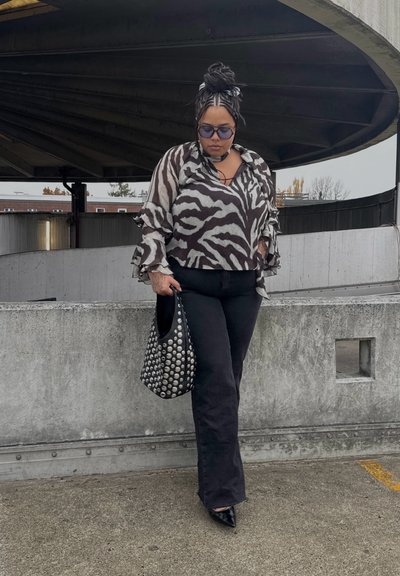 Blusa de estampado de cebra en blanco y negro con mangas con volantes, combinada con pantalones negros acampanados y un bolso negro con tachuelas, sobre un fondo de cemento.