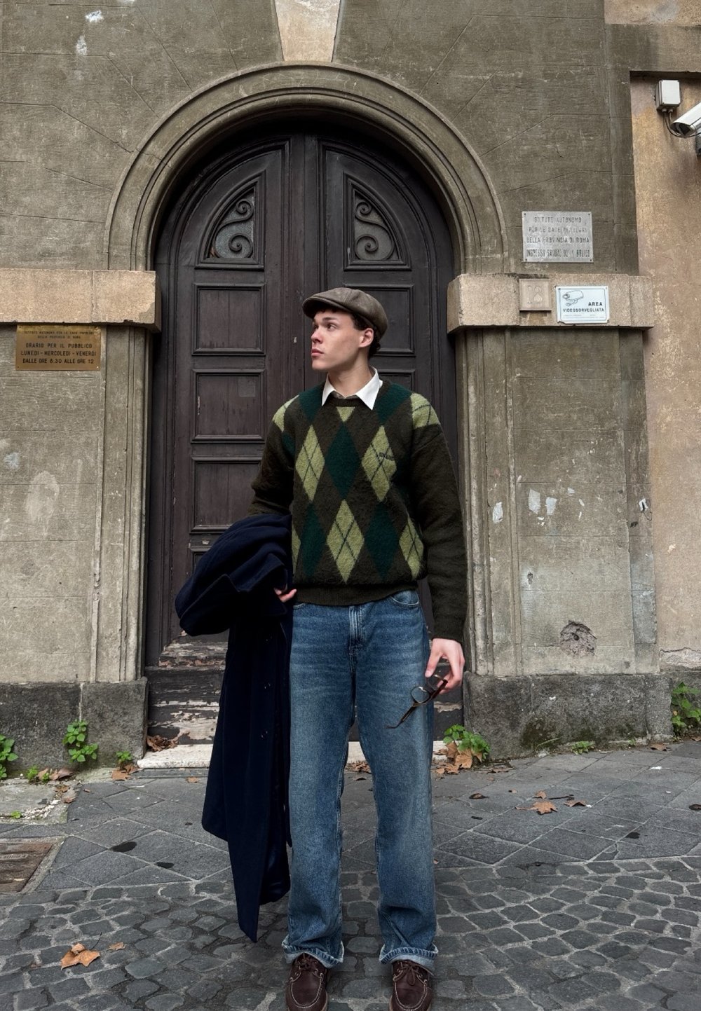 Un pull en argyle vert et marron sur une chemise blanche, un jean bleu, des chaussures marron, tenant des lunettes, avec un manteau noir drapé sur un bras.
