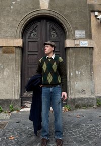 Un pull en argyle vert et marron sur une chemise blanche, un jean bleu, des chaussures marron, tenant des lunettes, avec un manteau noir drapé sur un bras.