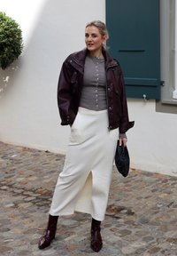 Bordeaux leren jack, grijze knooptop, crèmekleurige midi-jurk met zijsplit en bordeauxkleurige puntige laarzen. Zwarte gestructureerde clutch. Klinker achtergrond.