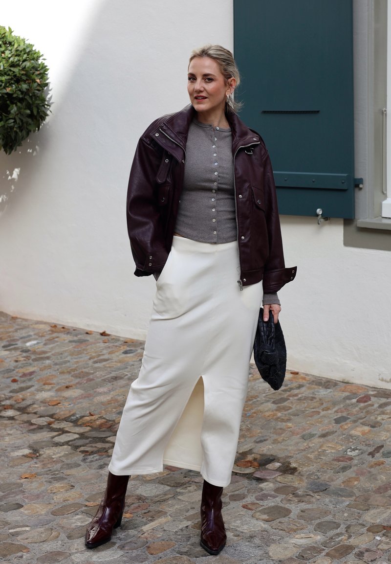 Veste en cuir bordeaux, haut gris à boutons, jupe midi crème avec fente sur le côté et bottes pointues bordeaux. Pochette noire texturée. Fond en pavés.